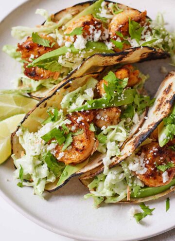 Three shrimp tacos on a plate with lime wedges beside them.