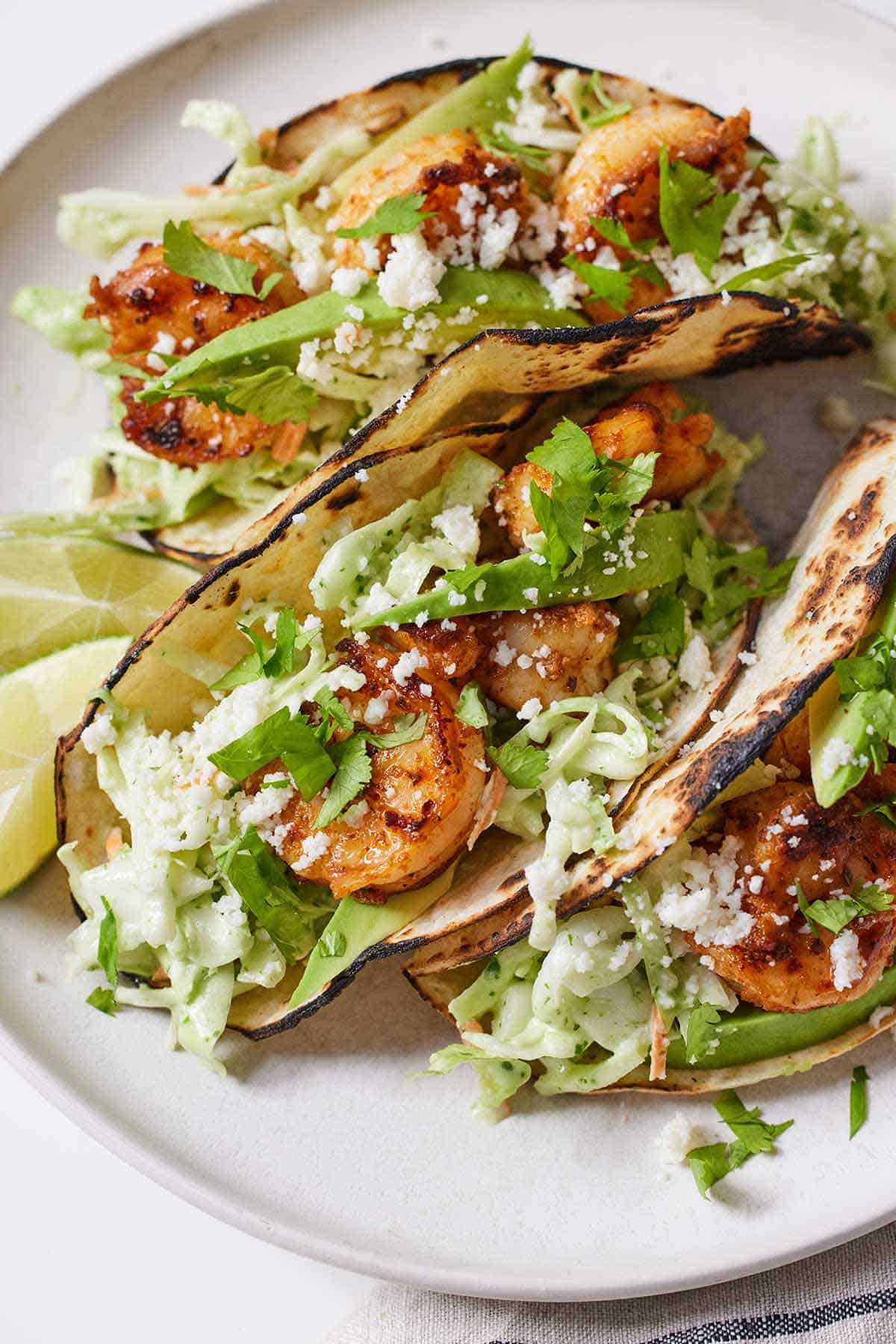 Three shrimp tacos on a plate with lime wedges beside them.