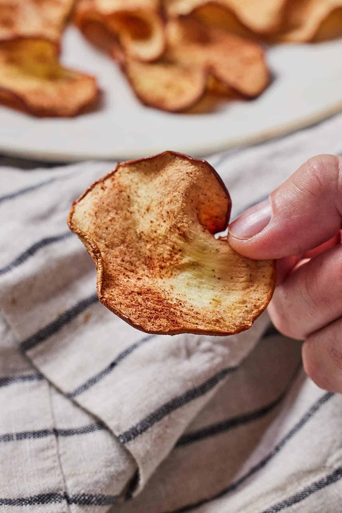 A hand holding up an air fryer apple chip in front of a plate of more chips.