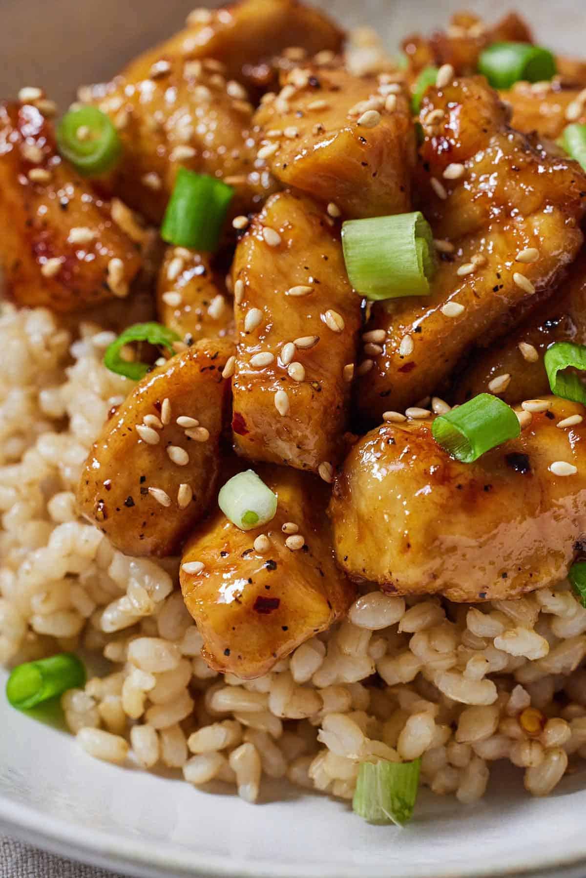 Close up of honey garlic chicken bites over brown rice with sesame seeds and green onions on top.