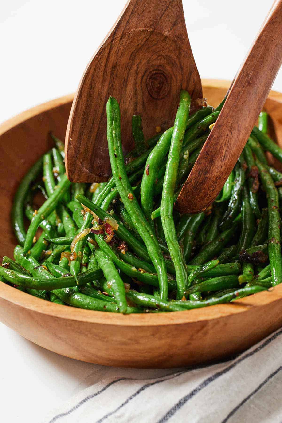 A bowl of crispy shallot green beans with wooden serving spoons tossing them.