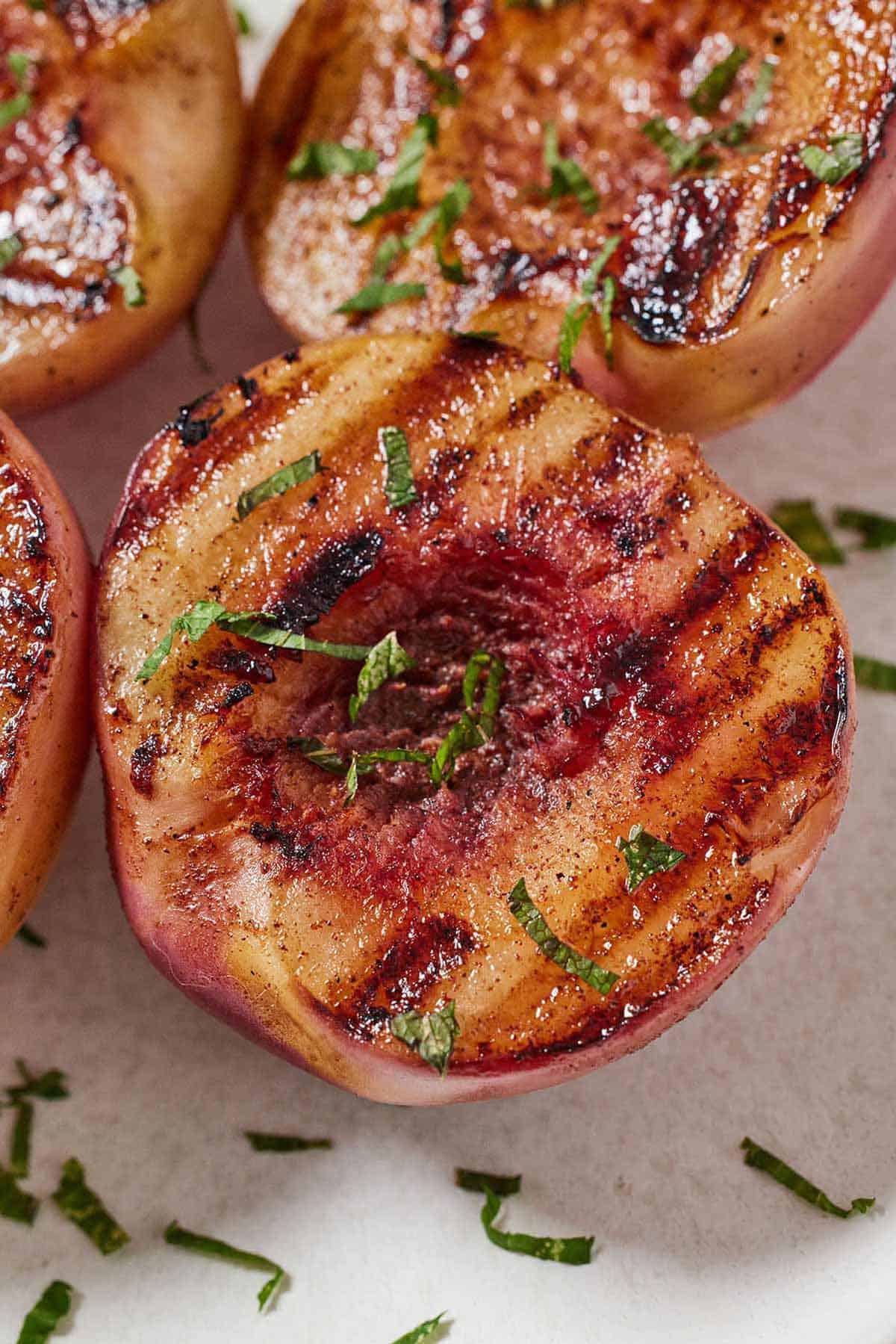 Close up of a grilled peach with more in the background with fresh chopped mint on top.