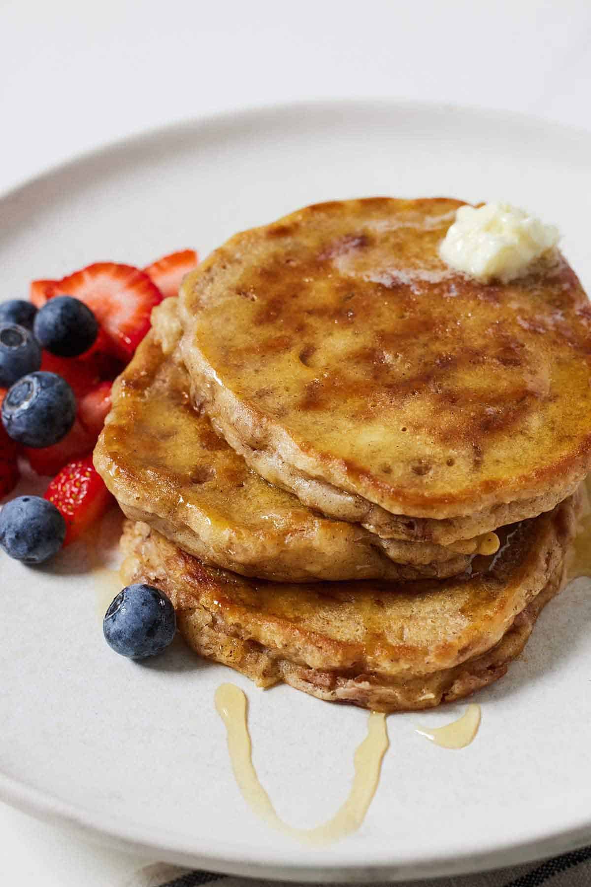 A plate with three apple pancakes stacked on top of each other with fresh berries on the side and syrup drizzled over top.