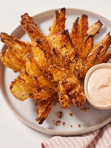 An air fryer blooming onion on a white plate with a dipping sauce on the side.