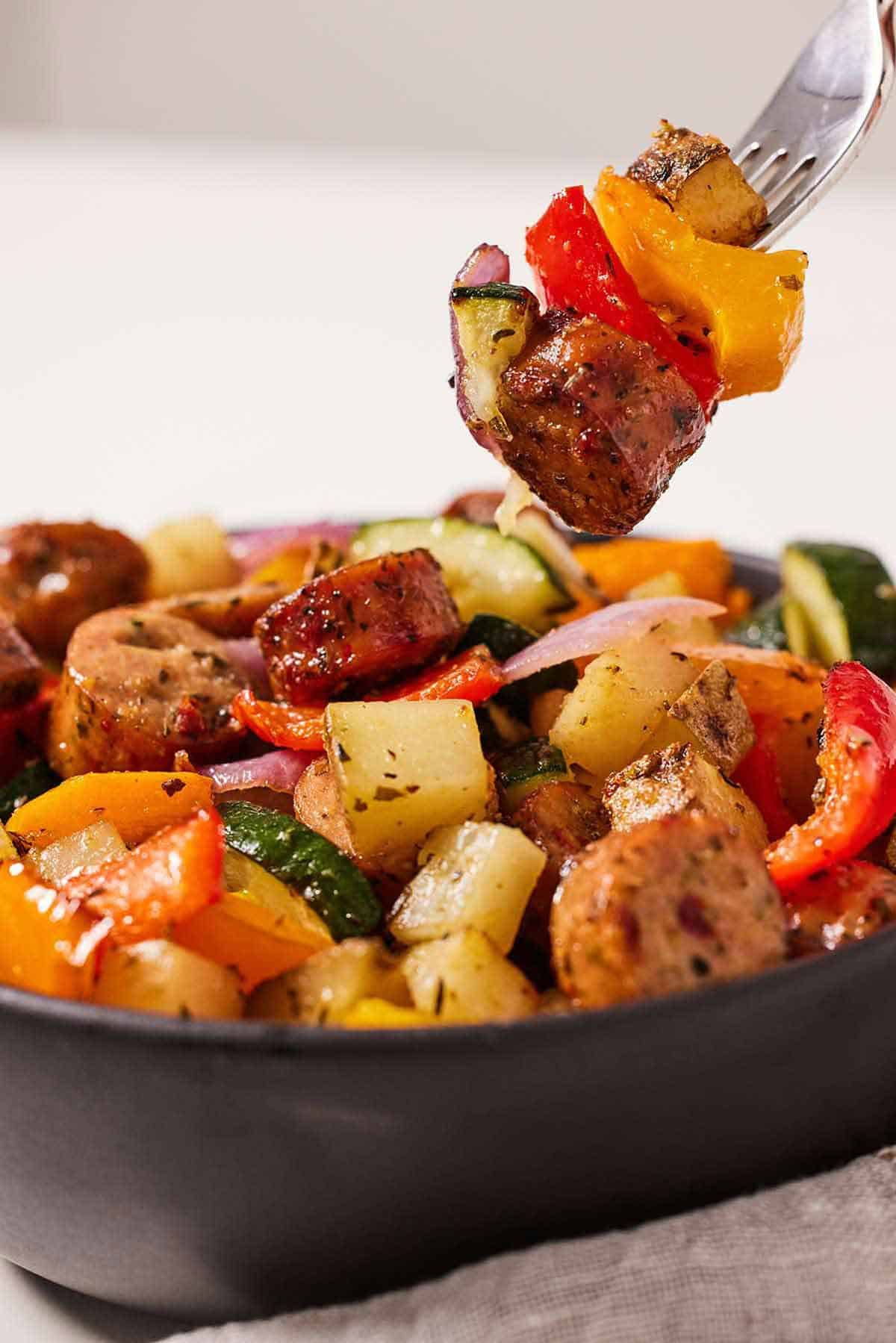 A fork with a bite of air fryer sausage and vegetables being lifted from a bowlful. .